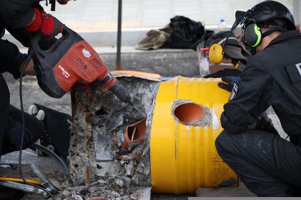 Mit einem Pressluftbohrer wird ein gelbes Fass vearveutetm ab dem noch eine Person mit Kopfhörern sitzt. Der offen liegende Beton und andere Dinge im Fass wie rostige Nägel sind zu sehen, auch ein Steg in einem Plastikrohr, eine Ankettvorrichtung.