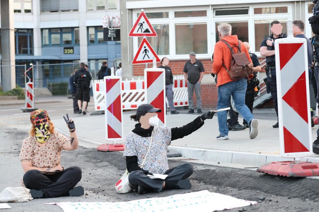 Zwei Personen sitzen auf der Straße, die rechte Person hält den rechts auf dem Bürgersteig vorbei laufenden Menschen ein Flugblatt hin. Menschen laufen auf dem Bürgersteig auf das Firmengelände, zwischen Baustellenbegrenzungen.