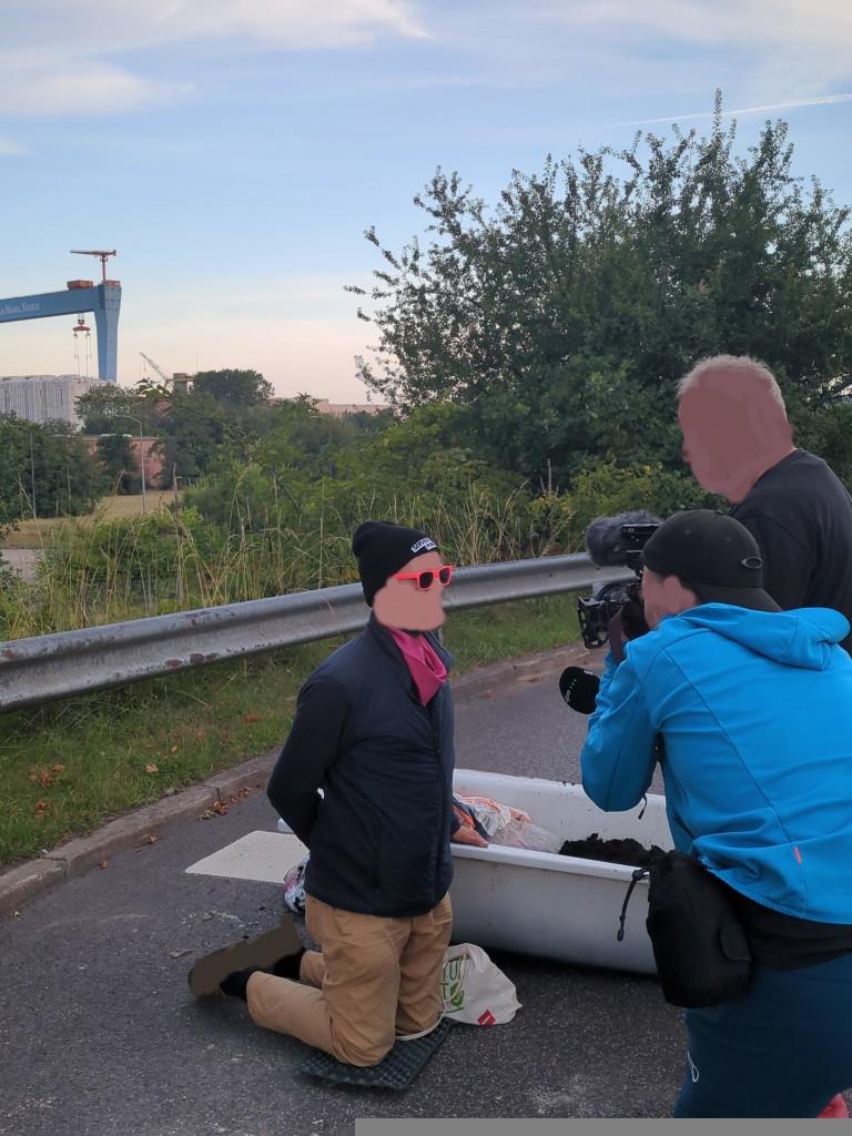 Eine Person mit der Hand in der Badewanne wird interviewt, eine andere Person hält eine Kamera, eine weitere ein Mikrofon. Im Hintergrund ist der charakteristische Kran von TKMS zu sehen.