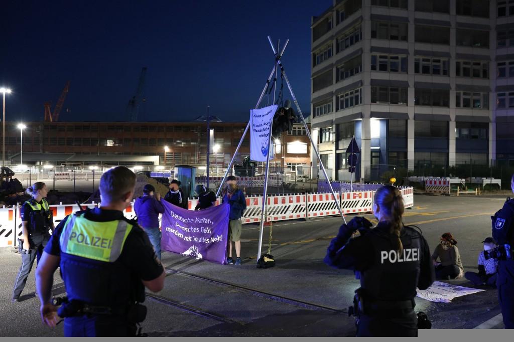 Es ist noch dunkel, es gibt Lichter vor einigen Gebäuden. Vor einem aufgebauten Tripod (dreibeiniges Gestell), in dem eine Person und ein Transparent hängen, stehen Menschen mit einem lila Transparent auf dem steht "Deutsche Waffen, deutsches Geld, morden mit in aller Welt" - einige Polizisten bewegen sich aus dem Vordergrund auf sie zu,
