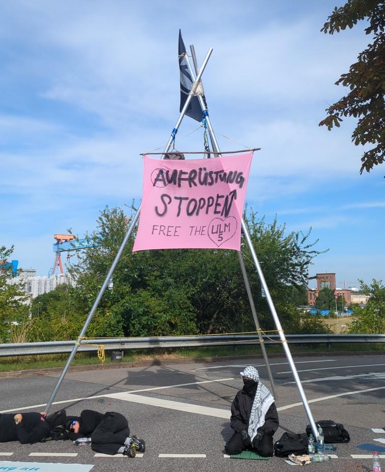 Ein Mensch sitzt, andere liegen unter einem dreibeinigen Gestell auf einer Straße an dem ein rosa Transparent mit der Aufschrift "Aufrüstung stoppen - free the ulm 5" steht.