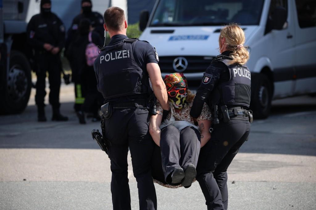 Eine Person wird von zwei Polizist*innen weggetragen, im Hintergrund wartet eine Polizeifahrzeug.